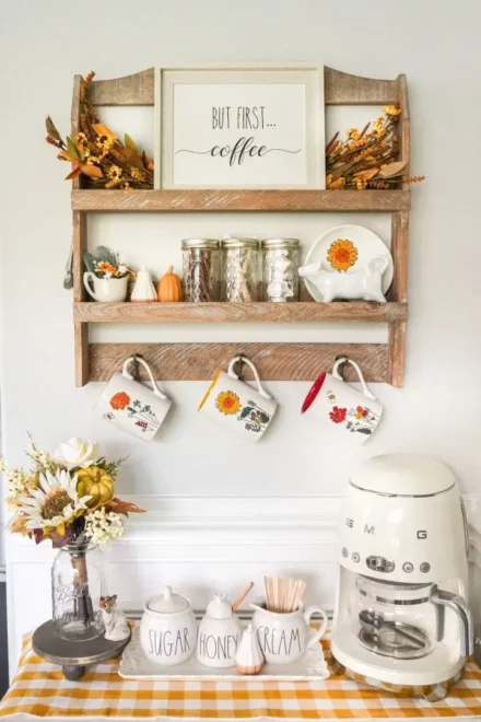 Farmhouse coffee bar with a white coffee machine, jars labeled sugar, honey, and cream, a yellow gingham cloth, and hanging mugs under a wooden shelf. The shelf displays mason jars, fall decor, and a "But First, Coffee" sign.