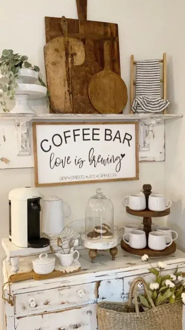 Rustic farmhouse coffee bar with a white coffee maker, cups on a wooden tiered tray, and a sign reading "COFFEE BAR love is brewing". Decor includes wooden cutting boards, striped towel, and a floral arrangement.