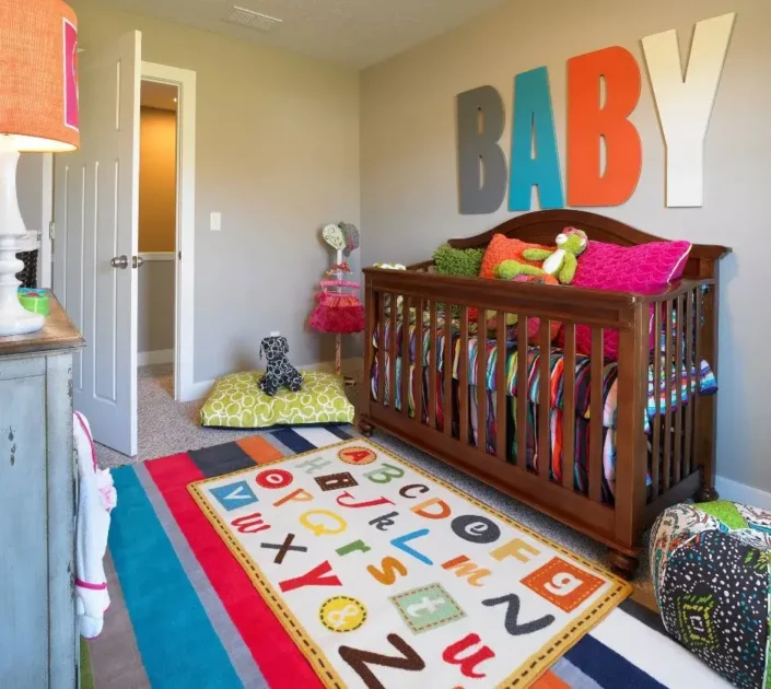 A brightly decorated nursery with a wooden crib filled with colorful pillows and stuffed animals. Large letters spelling "BABY" are mounted on the wall above the crib. The room features a vibrant alphabet-themed rug on the floor, a lamp with an orange shade on a dresser, and various toys and cushions scattered around. The door to the room is open, revealing a hallway with beige carpeting.