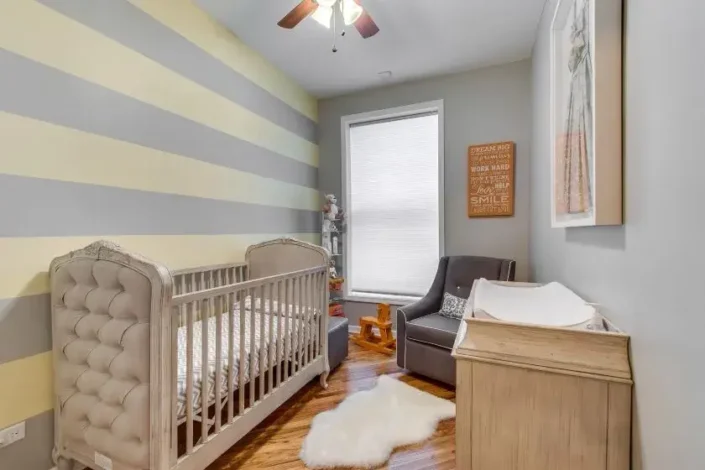 Gender-neutral nursery with a tufted crib, comfy armchair, changing table, and bookshelf. Light yellow and gray striped walls, white rug, and motivational sign