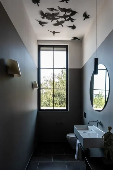 A modern bathroom with gray walls, featuring a tall window providing natural light and a view of greenery outside. The ceiling is adorned with a flock of bird-shaped decorations in flight formation. The room includes a white sink, wall-mounted faucet, mirror, and minimalistic light fixtures.