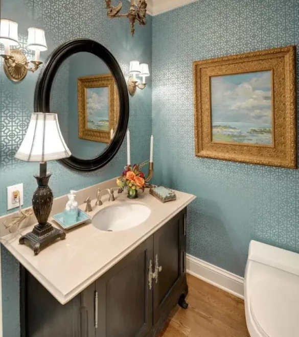 A bathroom with blue wallpaper, white vanity with built-in sink, oval mirror, seascape pictures, and a table lamp.