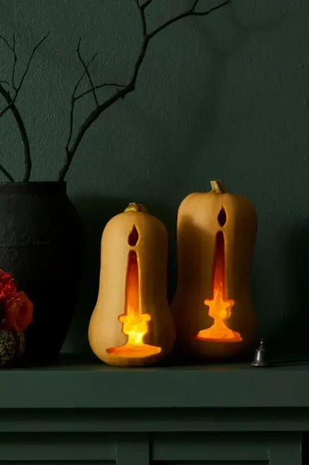 Butternut squashes carved like jack-o’-lanterns with lit candles inside, accompanied by a black vase with branches and an orange flower, perfect for easy and creative Halloween decorations.