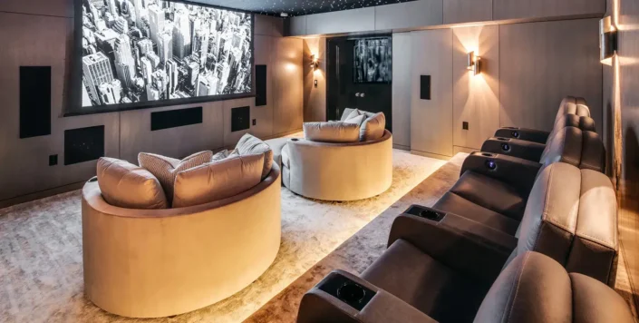 Modern home theater room with a large screen displaying a black and white movie scene, multiple rows of seating including leather recliners and round sofas, and ambient lighting fixtures, creating an inviting atmosphere perfect for basement movie theater