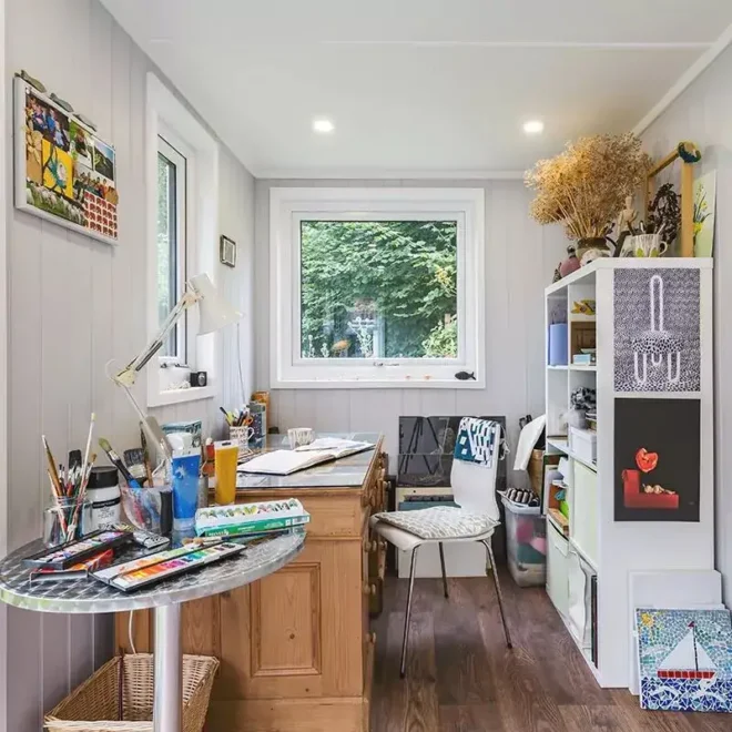 A brightly lit art studio with a wooden desk, art supplies, a white chair, and a shelving unit filled with books and decorative items. Windows provide natural light and a view of greenery.