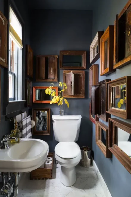 A bathroom with dark blue walls, white pedestal sink, asymmetrical arrangement of wooden frames, and a vase with yellow flowers.