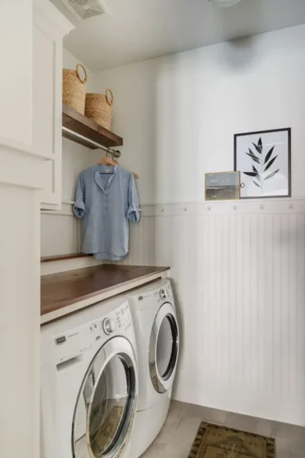 Laundry room with two white front-loading washing machines, wooden countertop, wicker baskets, shelves with folded items, and a framed plant print.