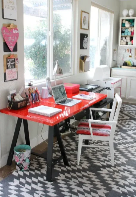 A home office with a red desk, white chair, and neatly arranged office supplies. The desk is in front of a window, allowing natural light to flood the space.