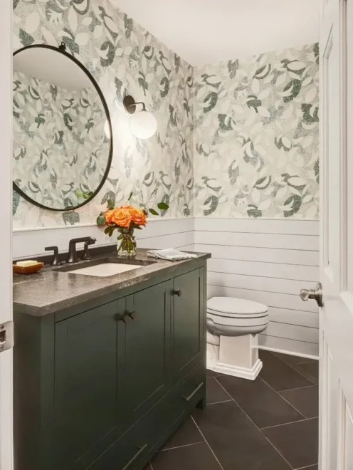 A bathroom with a dark green vanity, white countertop, round mirror, leafy wallpaper, and dark tiled floor.