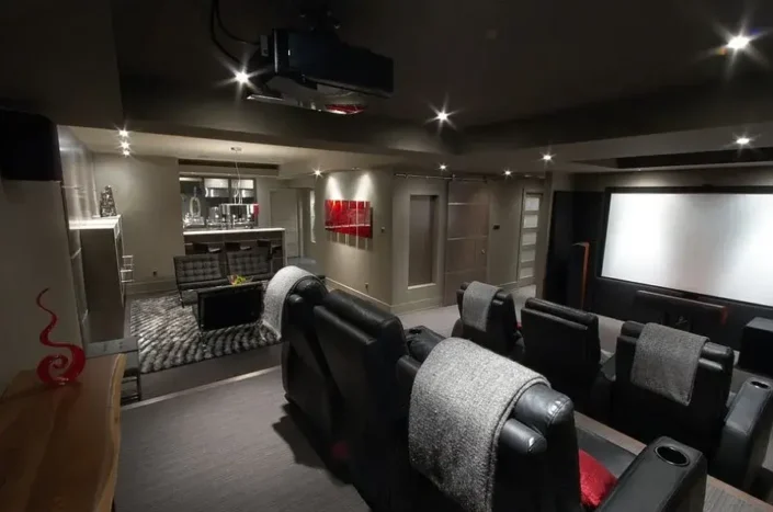 Home theater room with dark walls and ceiling, large screen, tiered plush armchairs, ambient wall-mounted lights, ceiling-mounted projector, and a counter with a sink and cabinets, perfect for basement home theater