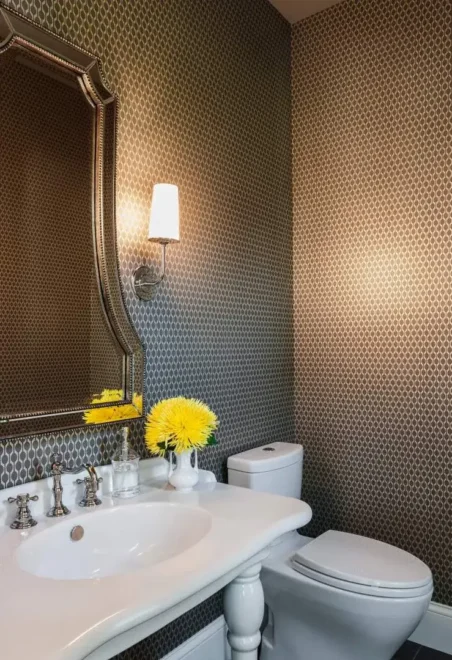 A bathroom with patterned wallpaper, white porcelain sink, ornate mirror, and yellow flowers in a vase.