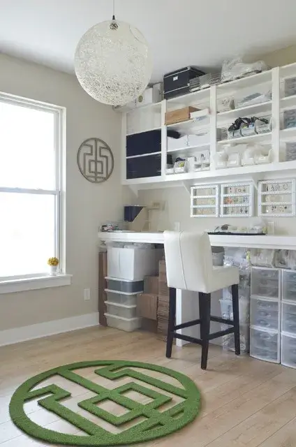 A well-organized craft or hobby room with a large work desk, white chair, and multiple shelves filled with crafting supplies. Natural light comes through a large window, highlighting a green circular rug and a decorative wall piece.