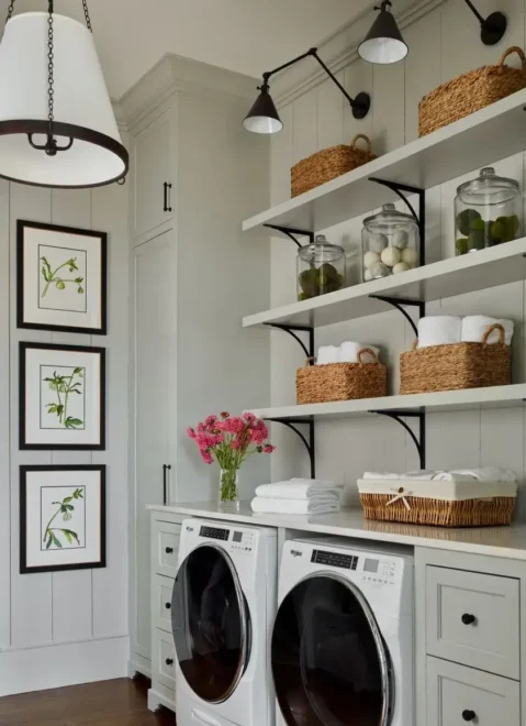 Laundry room with white front-loading washing machines, a white countertop, shelves with glass jars and woven baskets, and framed botanical illustrations on the wall.