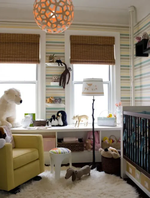 Gender-neutral nursery with a yellow armchair, geometric light fixture, stuffed animals, and striped wallpaper.