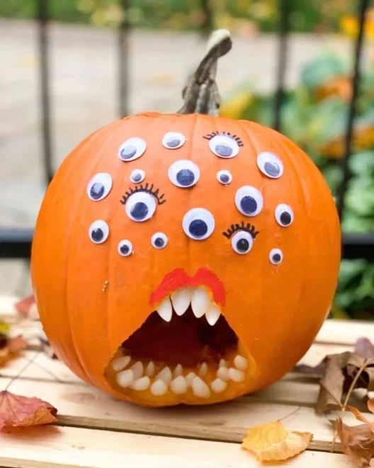 Pumpkin decorated with multiple googly eyes and a carved mouth with jagged teeth and a red tongue, surrounded by dry leaves, perfect for easy and humorous Halloween decorations