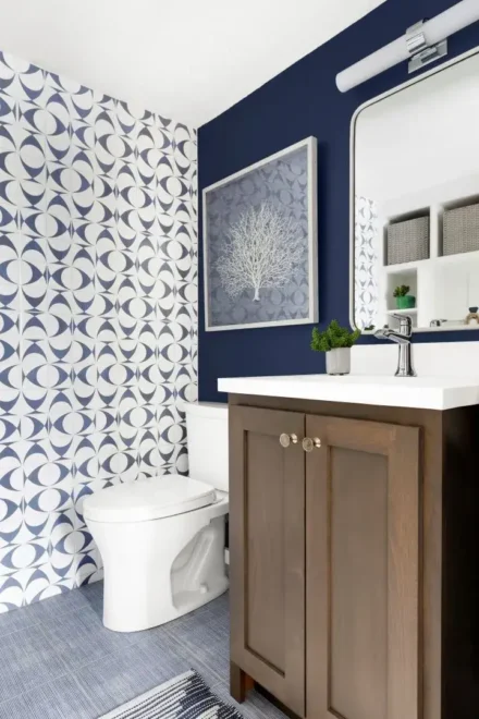 A contemporary bathroom with blue and white patterned wallpaper, dark blue wall, wooden cabinet under white sink, framed artwork above toilet, and striped rug on floor.