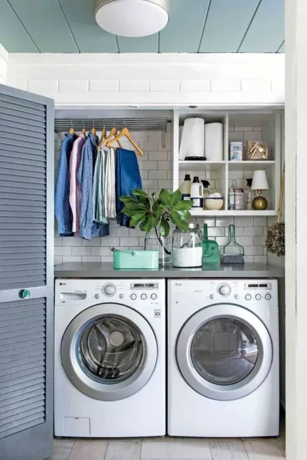 An interior laundry space featuring a front-loading washer and dryer side by side. Above the appliances, a white shelf holds towels, jars, and a plant. The wall behind is tiled with white subway tiles, and clothes are hanging on hangers to the left of the machines.