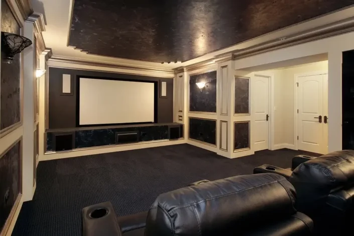 Home theater room with dark walls and ceiling, plush carpeted floor, large projection screen, black leather reclining chairs with cup holders, and built-in cabinetry, ideal for basement home theater room