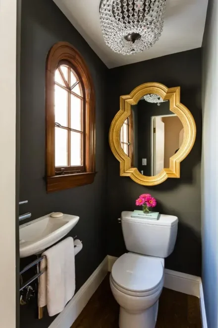 A small bathroom with dark-colored walls, featuring a white toilet with a small wooden shelf above it holding a pink flower arrangement. To the left of the toilet is a white sink with no visible cabinetry, and above it hangs an oval-shaped mirror with an ornate gold frame. An arched window with a brown wooden frame allows natural light into the space. The ceiling has an elaborate chandelier-style light fixture.