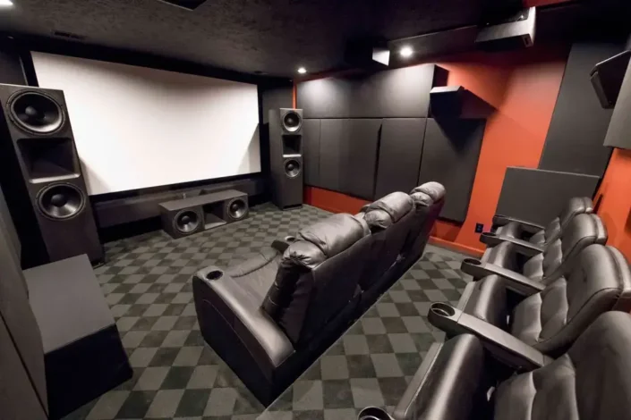 Home theater room with a large screen flanked by two large speakers, multiple rows of black leather reclining seats, acoustic panels on the walls, and patterned carpeting, creating a high-end cinema experience perfect for basement media room design