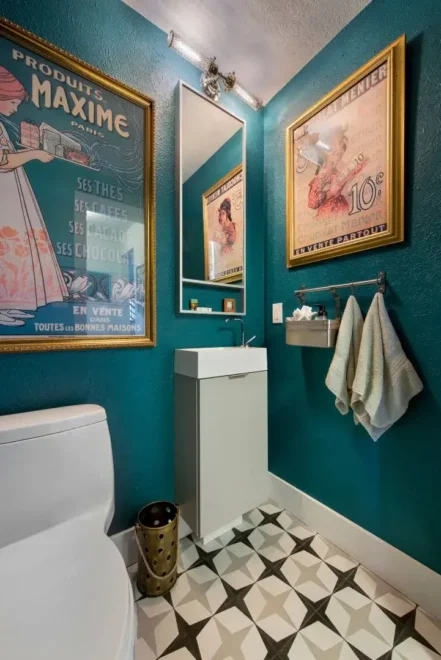 A stylish bathroom with teal walls, vintage French posters, white toilet and sink cabinet, geometric patterned floor tiles, wall-mounted light fixtures, and towels on a rack.