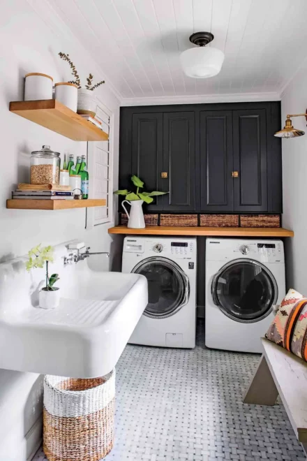 An interior laundry room with a white utility sink on the left, a wicker basket underneath, and a silver faucet. On the right, there are two front-loading laundry machines with a wooden countertop above them. Black cabinets with brass handles are mounted above the countertop, which holds jars, bottles, and a potted plant. The floor is tiled in a geometric pattern with shades of gray and white.