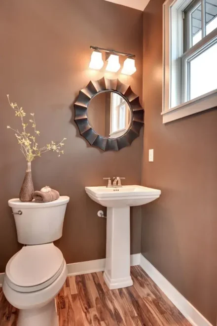 A small bathroom with brown walls, white pedestal sink, ornate round mirror, white toilet, and a window with frosted glass.