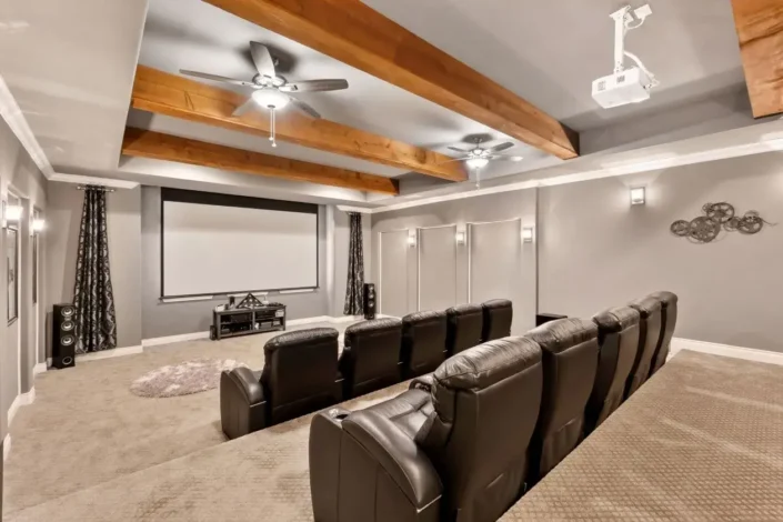 Home theater room with dark upholstered reclining chairs in two rows, light-colored carpeted flooring, gray walls, dark curtains, wooden ceiling beams, ceiling fans, and a mounted projector, perfect for finished basement movie room