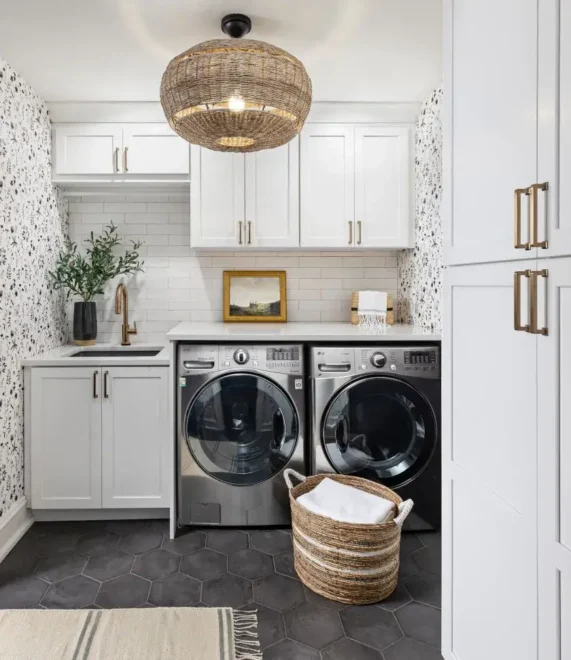 A modern laundry room with white cabinetry and stainless steel front-loading washer and dryer. Above the appliances, there is a small framed picture between two cabinets. To the left, a potted plant sits next to a sink with a faucet on the countertop. The floor features dark hexagonal tiles with a patterned rug in front of the washer and dryer. A woven laundry basket filled with white linens is on the floor to the right. The wall has floral wallpaper, and a wicker light fixture hangs above.