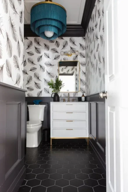 A bathroom with feather-patterned wallpaper, black hexagonal floor tiles, white sink cabinet, gold-framed mirror, and blue drum pendant light.