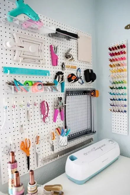 A craft room setup with a pegboard displaying various tools and supplies, including scissors, rulers, glue guns, and cutting tools, with a Cricut machine and thread organizer nearby.