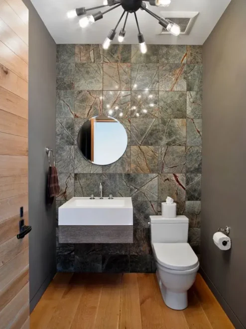 A modern bathroom with a wall-mounted sink, geometric stone tiles, round illuminated mirror, and light wooden floor.