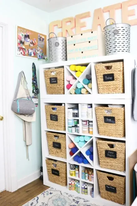 A well-organized craft storage area with labeled wicker baskets and paint supplies, neatly arranged on white shelving. Above the unit, large wooden letters spell out "CREATE," adding a decorative touch to the space.