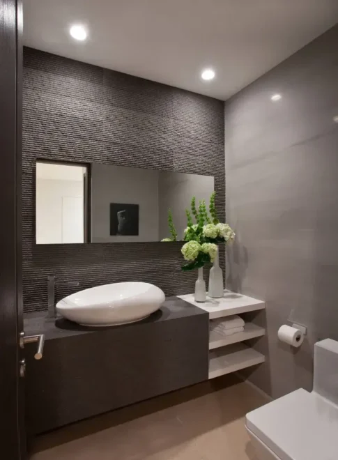 A modern bathroom with a dark brown vanity, white vessel sink, rectangular mirror, and small white stand with a vase of green plants and white flowers.