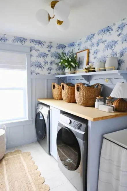 Laundry room with blue floral wallpaper, wooden countertops, grey appliances, and white cabinets with shelves holding baskets and decor.