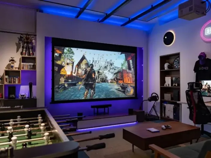 Modern gaming setup with ambient purple and blue lighting, featuring a large screen displaying a medieval-style video game, a gaming chair, a shelf with collectibles, and a foosball table, perfect for basement media room design