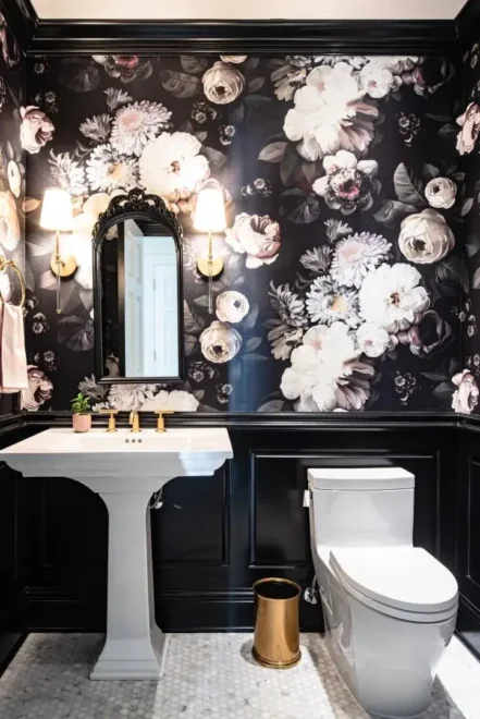 A bathroom with dark floral wallpaper, white pedestal sink with gold fixtures, ornate black-framed mirror, white toilet, and black tile flooring.