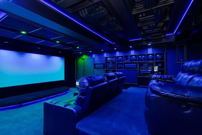 Home theater with tiered plush dark blue recliner chairs, ambient blue lighting, framed items on the walls, and a large projection screen, creating a high-end private cinema experience perfect for basement home theater