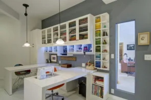 A modern, well-organized craft room with a large white desk and sewing machine, featuring white cabinetry with neatly folded fabrics and open shelves on the right.