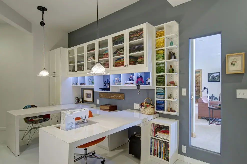 A modern, well-organized craft room with a large white desk and sewing machine, featuring white cabinetry with neatly folded fabrics and open shelves on the right.