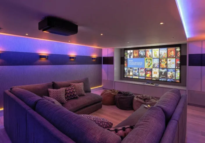 A modern home theater room with purple LED lighting, featuring a large screen displaying a selection of movies, a gray sectional sofa with patterned cushions, and a brown bean bag chair on the carpeted floor. The room includes wall-mounted speakers and a ceiling-mounted projector.